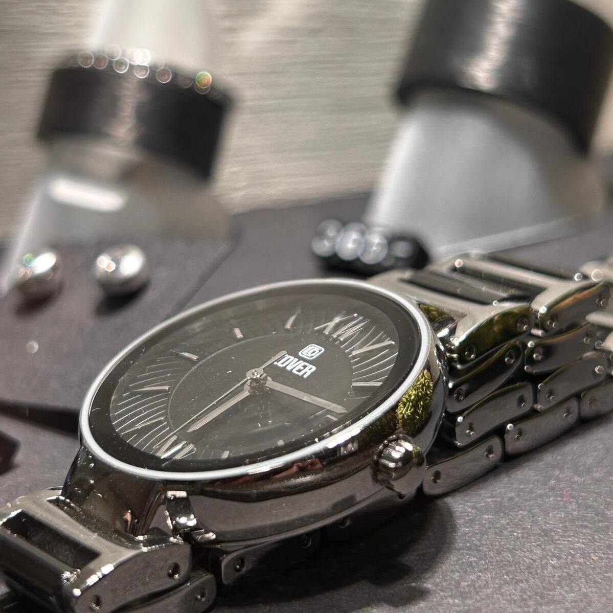 Uhr der Marke Cover, mit schwarzem Zifferblatt, im Hintergrund Fingerringe